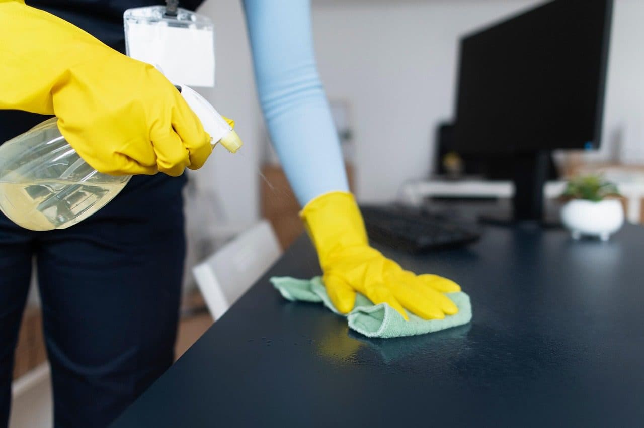 Reinigungskraft in dunkler Arbeitskleidung und gelben Handschuhen wischt einen dunklen Schreibtisch mit einem mintgrünen Mikrofasertuch ab, in der anderen Hand eine Sprühflasche. Im Hintergrund Monitor, Tastatur und eine kleine Pflanze.